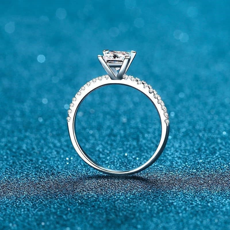 Princess cut on pavé band moissanite ring standing up on end on blue background